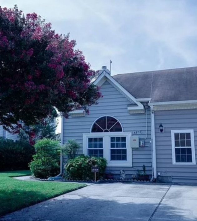 The exterior of The Achievable Dreams House — a single-story home with a crepe myrtle in bloom and a private driveway.